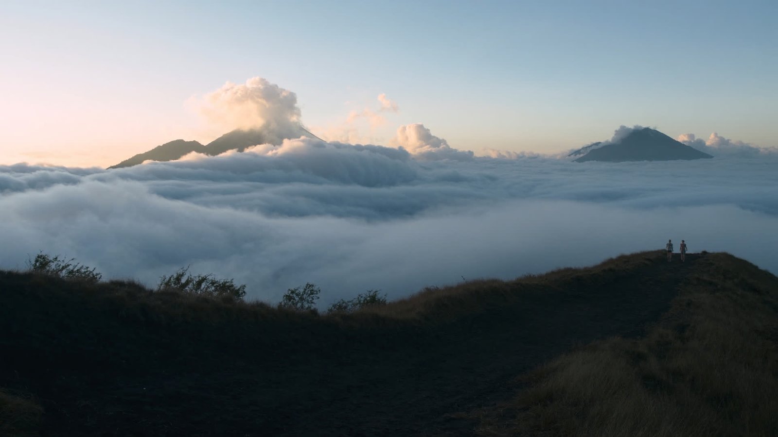 First light striking a sea of cloud from a volcanic ridge in Bali, with two distant figures on the rim.