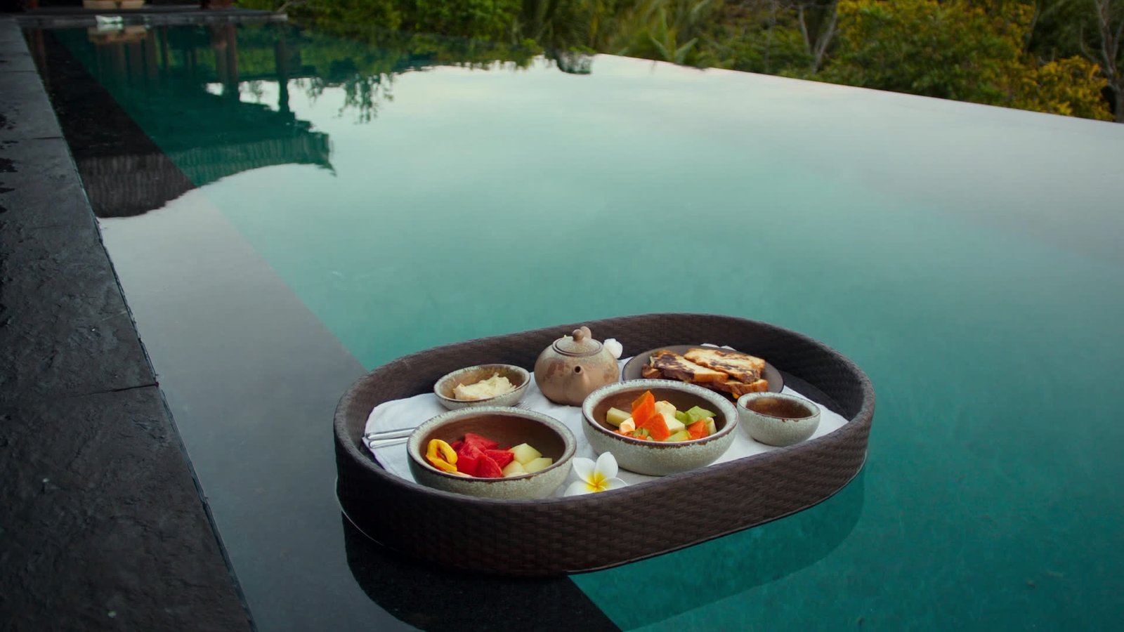 A rattan breakfast tray with ceramic bowls of fruit floating on a still infinity pool above a jungle valley.