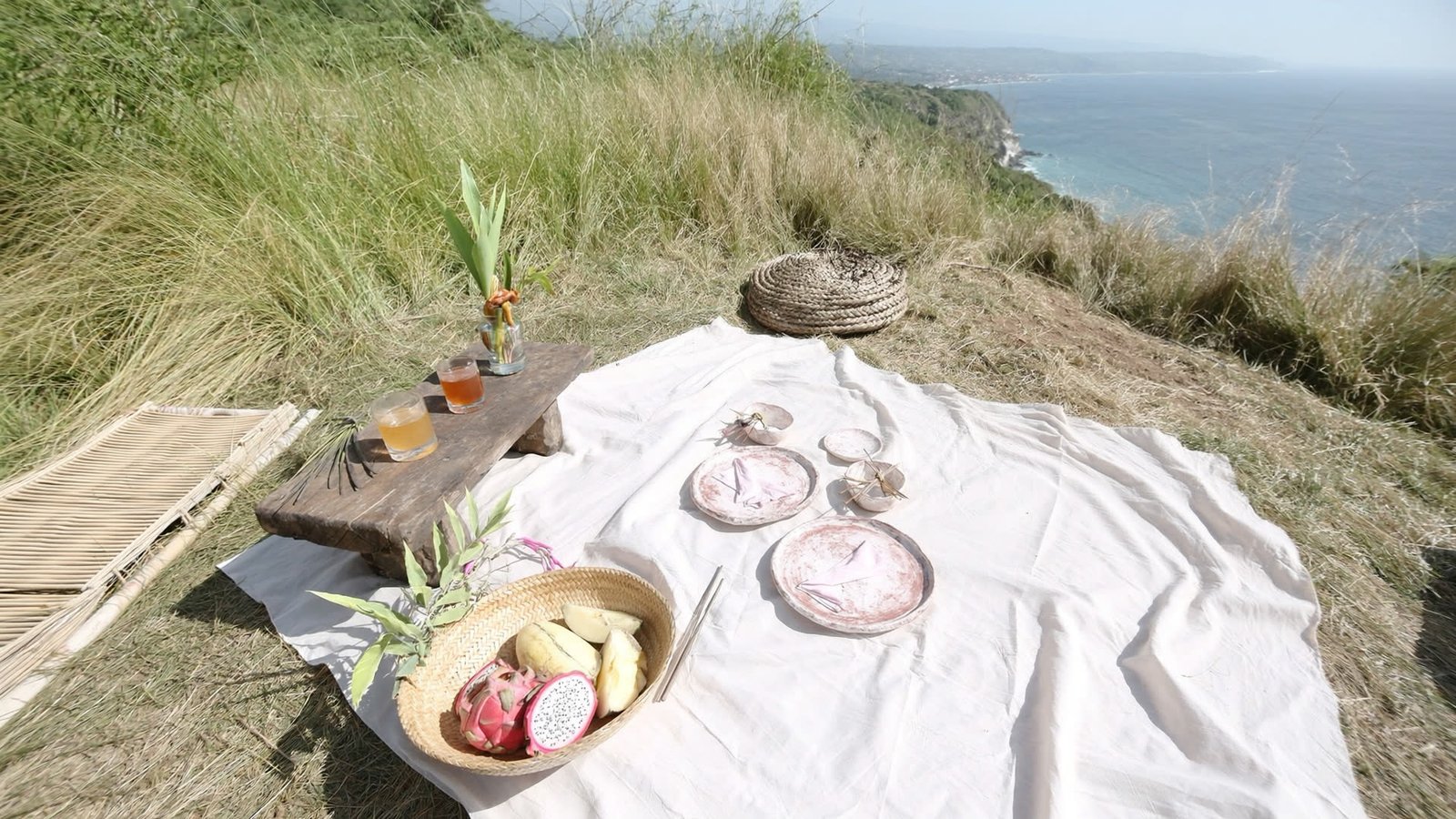 A picnic laid out on linen on a cliff meadow above the Bali coast, ocean to the horizon.