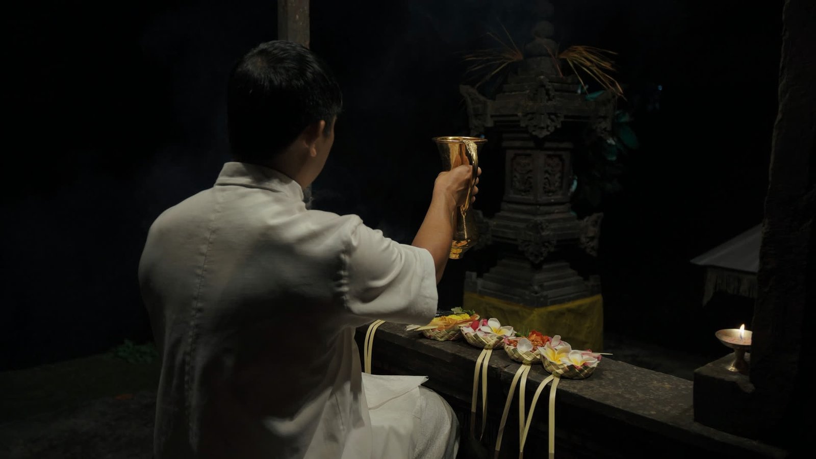 A Balinese priest in white performing a blessing at night, canang sari offerings and oil lamps on dark stone.