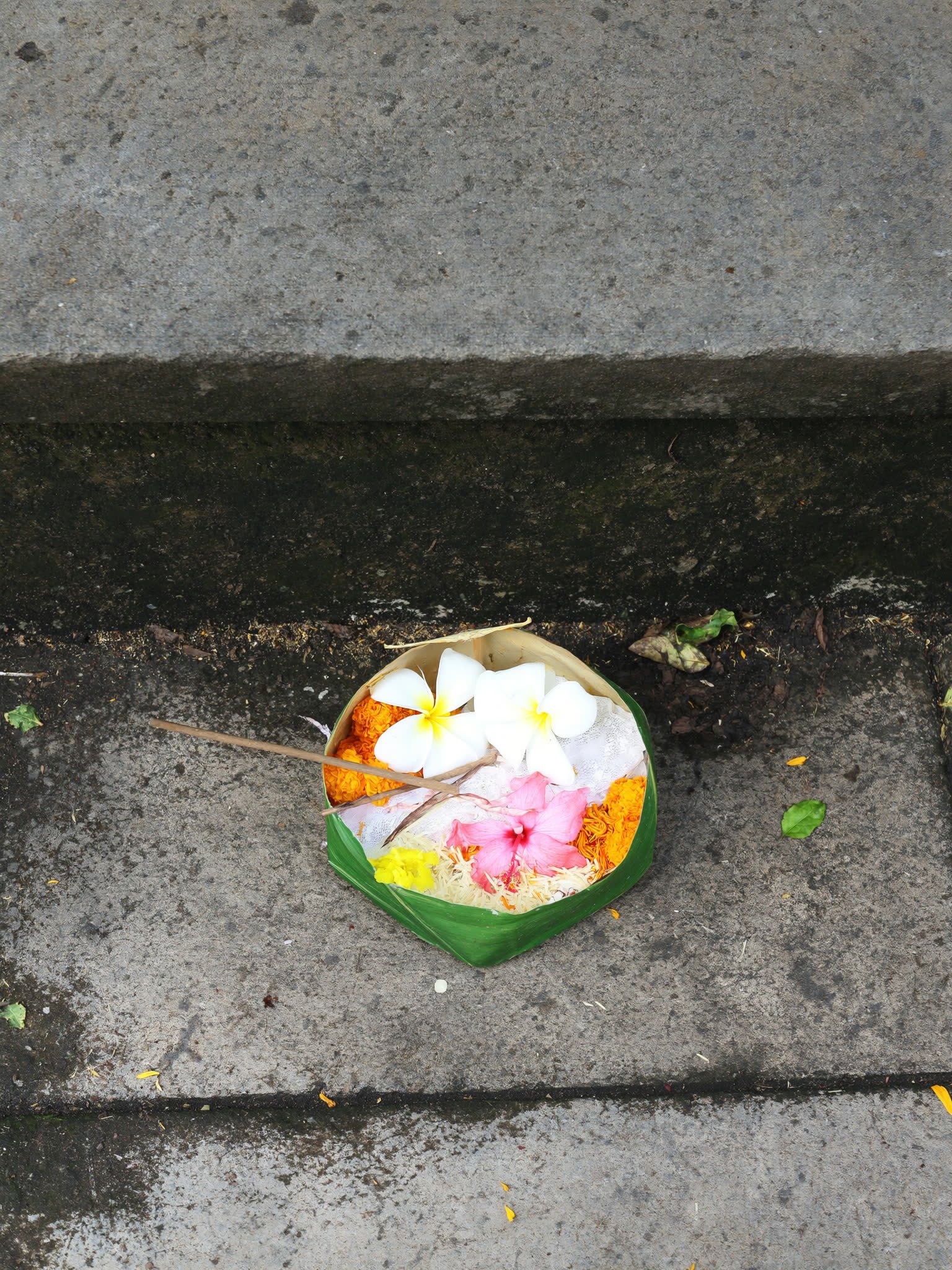 Two hands finishing a canang sari offering of folded coconut leaf filled with petals on a stone step.