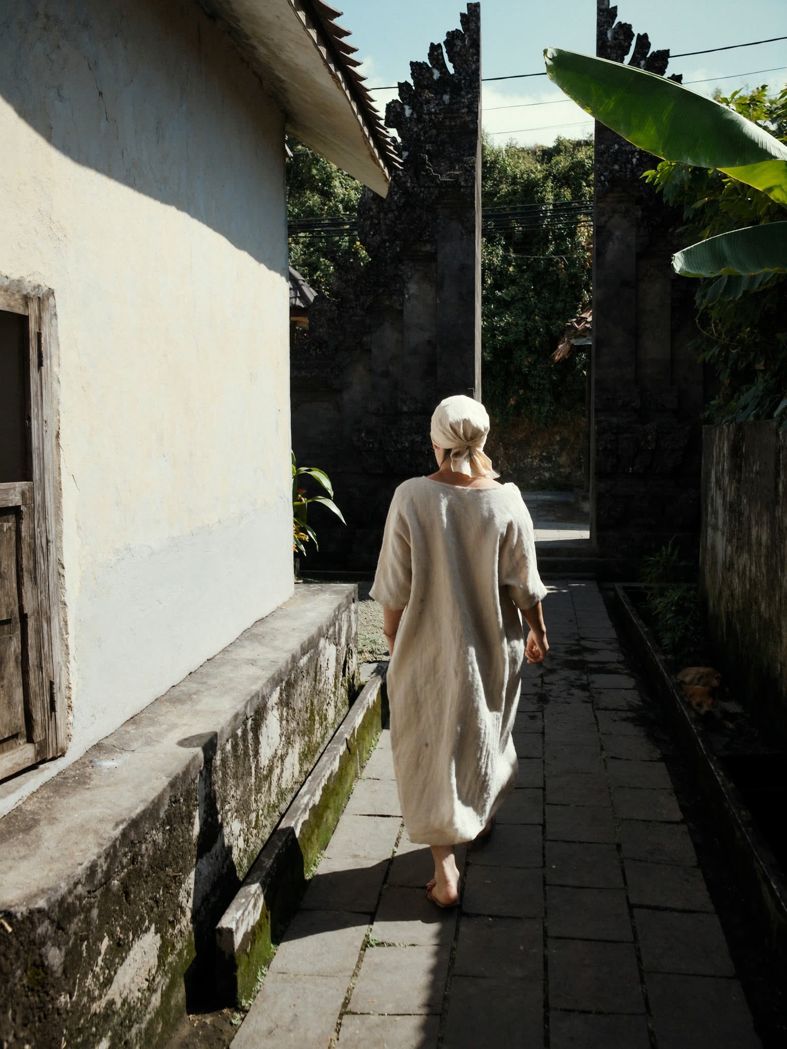 A woman in linen walking away up a narrow moss edged village lane toward a carved stone split gate.