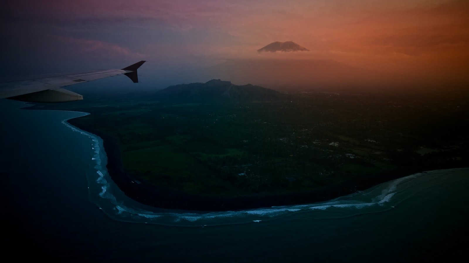 View from an aeroplane window on approach to Bali at dusk, surf lines along a dark beach below.